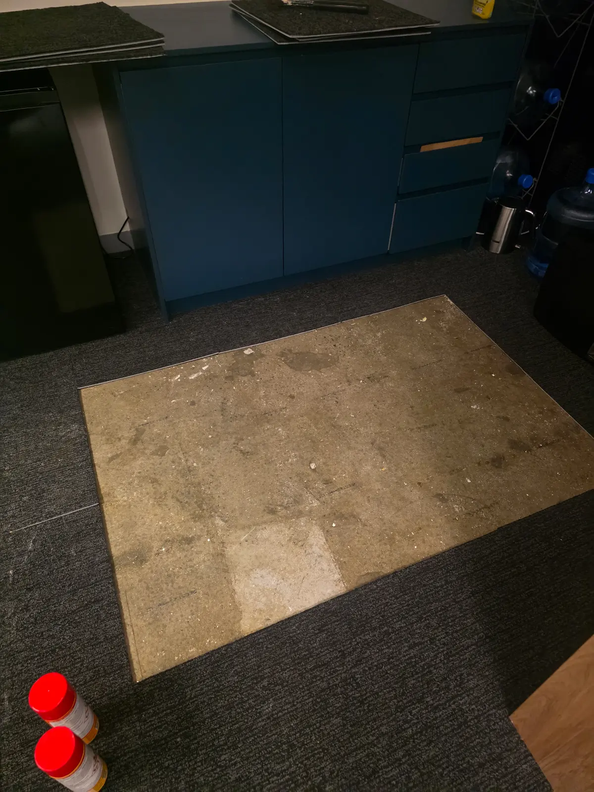 Carpet tiles lifted to expose a concrete floor slab for repair work in an office kitchen area